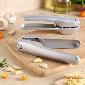 Two Zyliss Susi 4 Garlic Presses with ergonomic handles rest on a wooden cutting board surrounded by garlic cloves, cherry tomatoes, rosemary, and chopped garlic, making prep and cleanup easy with your favorite fresh ingredients.