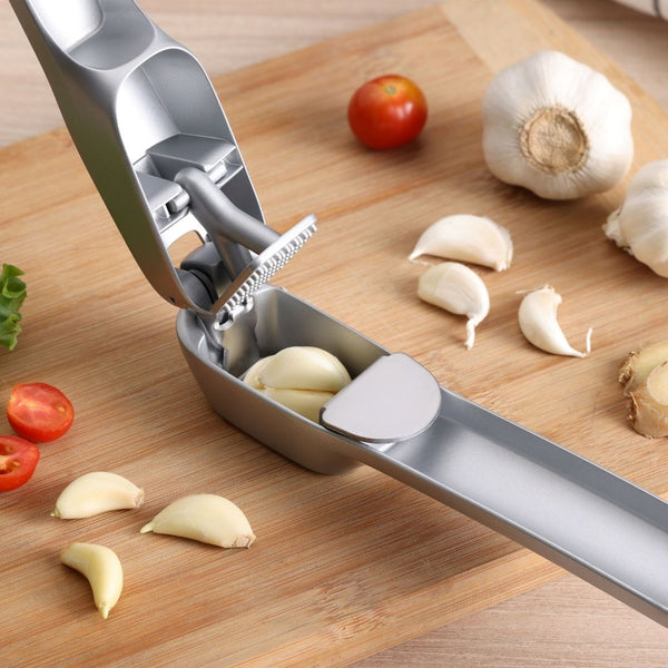 The Zyliss Susi 4 Garlic Press with ergonomic handles is shown with peeled garlic cloves, whole bulbs, and cherry tomatoes on a wooden cutting board for easy cleanup after prep.