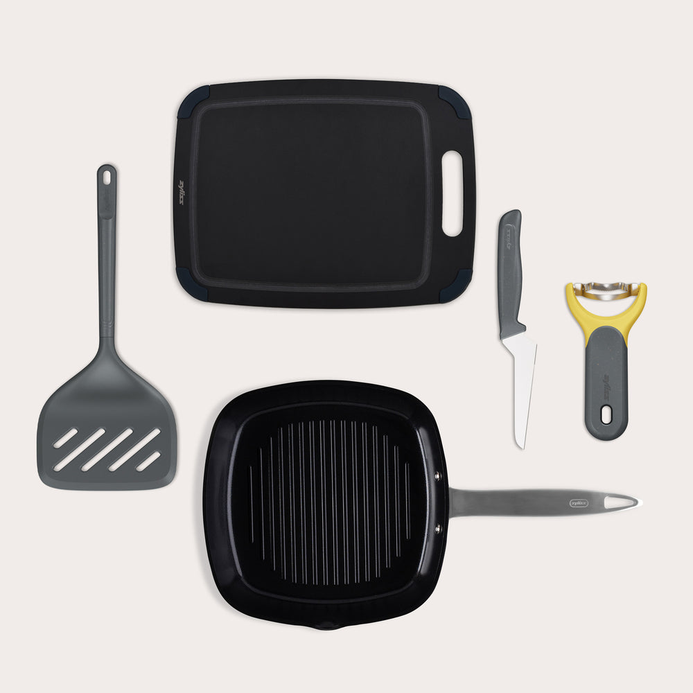 A flat lay of the Zyliss BBQ Bundle, featuring an Ultimate Pro Ceramic Grill Pan, XL Turner, black cutting board, grey-handled knife, and yellow vegetable peeler, all arranged on a light background.
