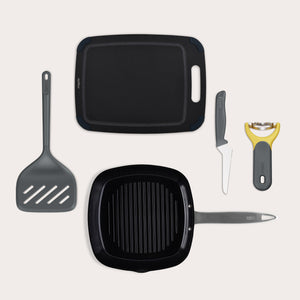 A flat lay of the Zyliss BBQ Bundle, featuring an Ultimate Pro Ceramic Grill Pan, XL Turner, black cutting board, grey-handled knife, and yellow vegetable peeler, all arranged on a light background.