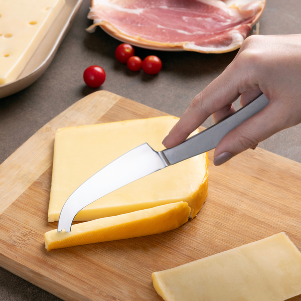 A hand slices a wedge of yellow cheese on a wooden cheese board using the Zyliss Signature Universal Cheese Knife. Sliced cheese, ham, cherry tomatoes, and Swiss cheese are visible in the background.