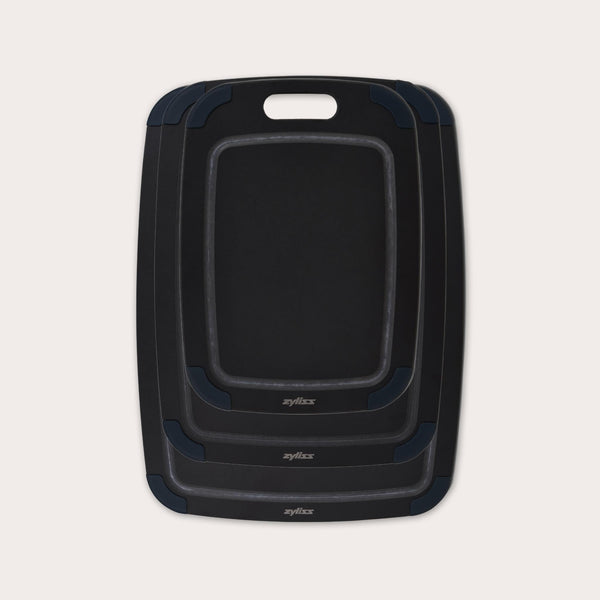 The Zyliss Wood Fibre Cutting Boards set features three nested black boards in different sizes, each with a handle slot and dark gray corner grips, made from eco-friendly recycled wood fibres and shown on a light background. #set of 3