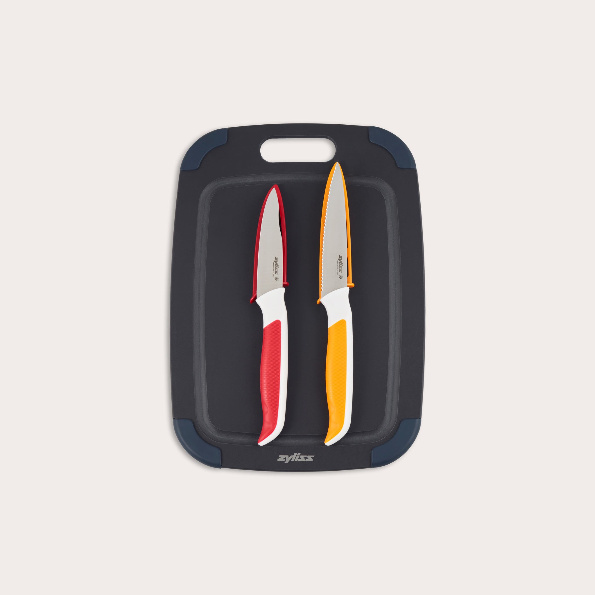 The Zyliss 3 Piece Wood Fibre Chopping Board & Knife Set includes a black cutting board with rounded corners and two knives—one red-handled and one orange-handled—both with silver blades. 