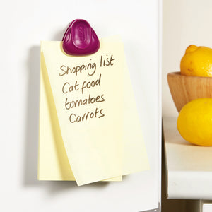 A yellow sticky note with a shopping list—cat food, tomatoes, carrots—is secured to a white fridge by a purple Zyliss Clip-All Bag Clip. Lemons rest in a wooden bowl on the counter nearby. An everyday kitchen essential. 