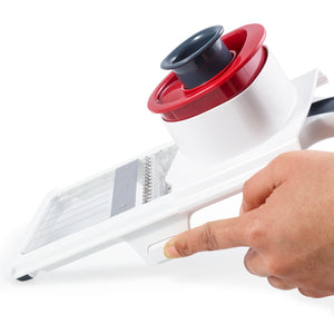A hand presses a button on the Zyliss Easy Control Handheld Slicer, which features an adjustable thickness setting, metal blade, and food holder. The slicer is white and red and is shown from a side angle against a white background.