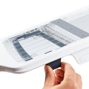 A hand pulls the black handle of the Zyliss Easy Control Handheld Slicer, a white slicer with adjustable thickness settings, metal blades, and a clear protective cover.