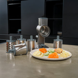 The Zyliss Drum Grater & 6 Drum Accessories rests on a kitchen counter. A plate in front displays sliced cucumber, grated cheese, carrot, and potato dishes, while sleek black cabinets add a modern touch to the kitchen setting.