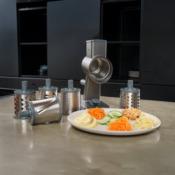 The Zyliss Drum Grater & 6 Drum Accessories rests on a kitchen counter. A plate in front displays sliced cucumber, grated cheese, carrot, and potato dishes, while sleek black cabinets add a modern touch to the kitchen setting.