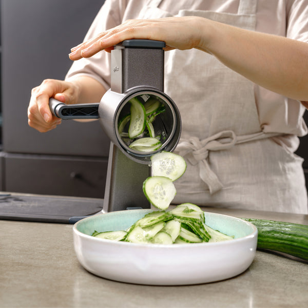 Wearing a light-colored apron, a person uses the Zyliss Drum Grater & 6 Drum Accessories with stainless steel drums to slice cucumber into thin pieces that fall into a white bowl on the kitchen counter.