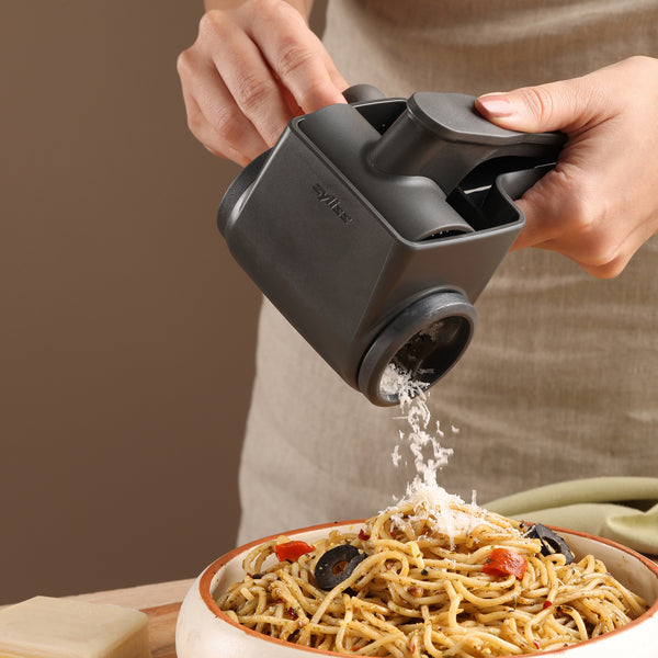 A person grates cheese onto pasta using the Zyliss Professional Rotary Cheese Grater, featuring a black handle and stainless steel drum. The pasta, garnished with olives and cherry tomatoes, sits in a cozy kitchen setting.