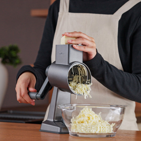 A person in a beige apron uses the Zyliss Gourmet Drum Grater to shred cheese into a glass bowl on a wooden countertop.