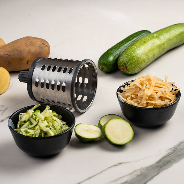 The Zyliss Gourmet Drum Grater is shown with shredded zucchini and potatoes in black bowls, alongside fresh zucchini slices, whole potatoes, and whole zucchini on a white marble surface.