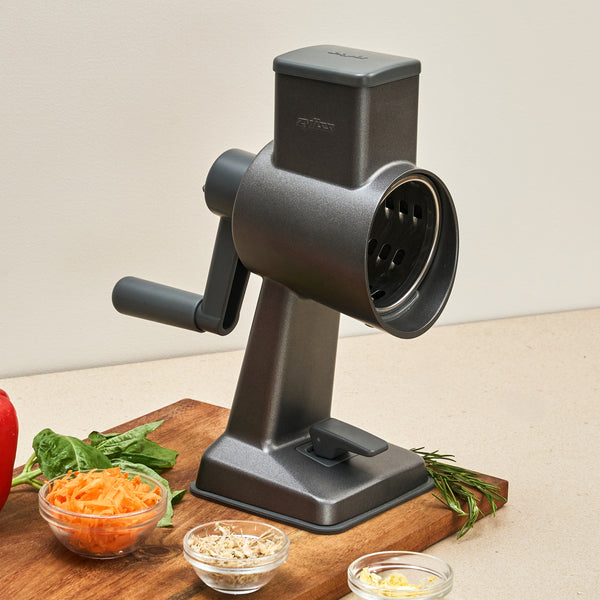 The Zyliss Gourmet Drum Grater sits on a kitchen counter beside a board with grated carrots and bowls of cheese and herbs—a stainless steel grater designed for effortless meal prep.