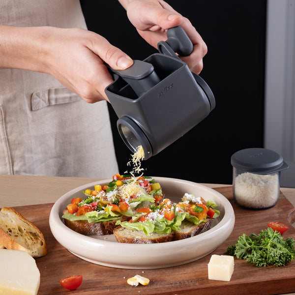 Using the Zyliss Professional Rotary Cheese Grater Set with a detachable container, cheese is grated onto open-faced vegetable sandwiches. Bread, cheese, a herb sprig, and a jar of grated cheese sit nearby on the table.
