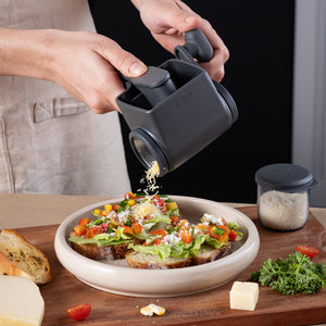 Using the Zyliss Cacio e pepe bundle, a person grates cheese with a black handheld rotary grater over veggie open-faced sandwiches, giving them a restaurant-quality finish. Bread slices, parsley, cheese, and grated cheese sit nearby.