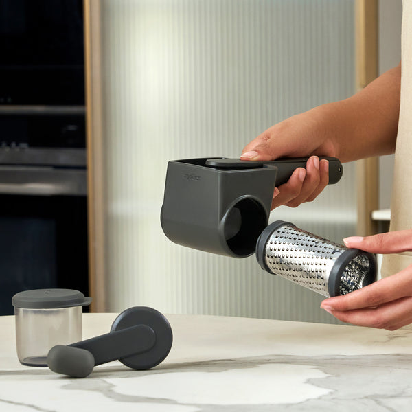 A person assembles the Zyliss Cacio e pepe bundle, a black rotary cheese grater, on the kitchen counter. Other parts like the container and plunger are nearby—perfect for grating cheese to top pasta dishes.