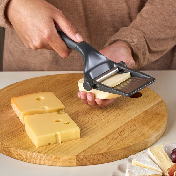 A person uses the Zyliss Dial & Slice Cheese Slicer, featuring an adjustable stainless steel blade, to cut cheese slices from a block on a round wooden cutting board while other cheese pieces are nearby.