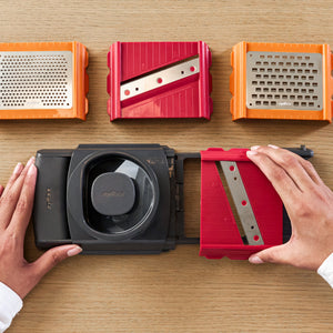 Hands attaching a red slicing blade to a black Zyliss 4-in-1 Slicer and Grater with Interchangeable Blades, with two additional grating plates and another slicer placed above on a wooden surface.