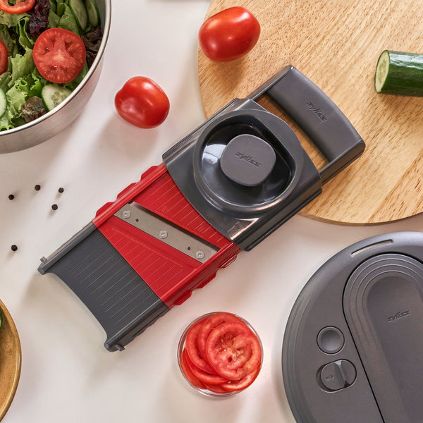 A Zyliss 4-in-1 Slicer and Grater with Interchangeable Blades surrounded by sliced tomatoes, a bowl of salad, and a cucumber on a kitchen surface.