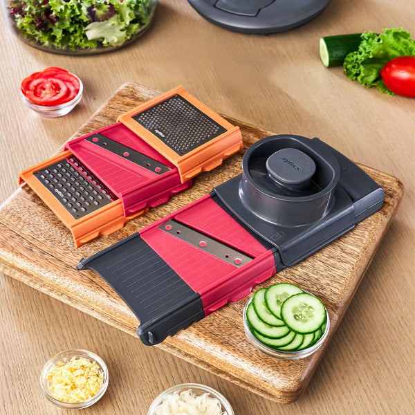 Two Zyliss 4-in-1 Slicer and Grater with Interchangeable Blades tools sit on a wooden cutting board next to bowls of sliced cucumbers, tomatoes, and shredded ingredients prepared with the compact kitchen tool.