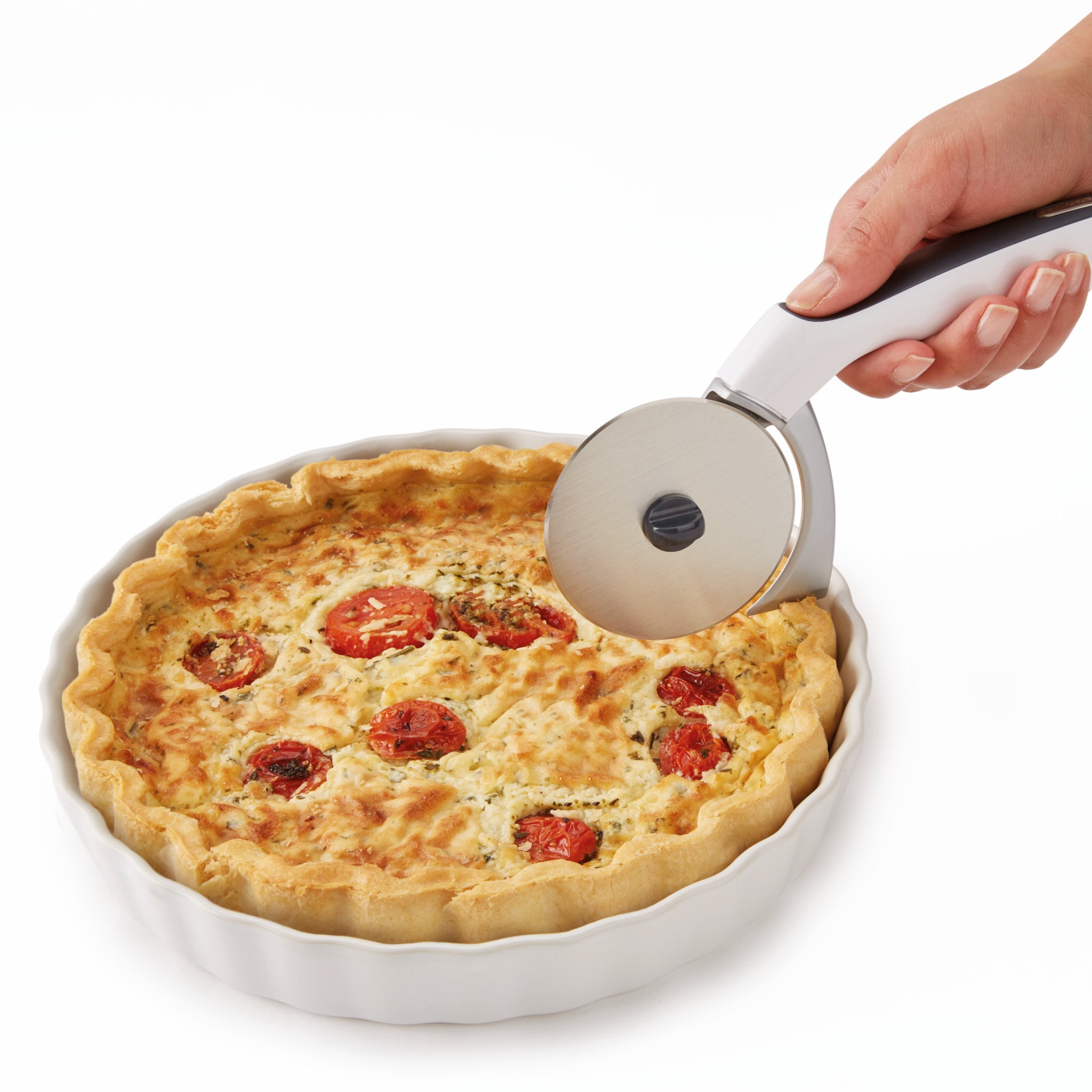 A hand holding a pizza cutter about to slice a baked quiche with tomatoes and herbs in a white ceramic dish on a white background.