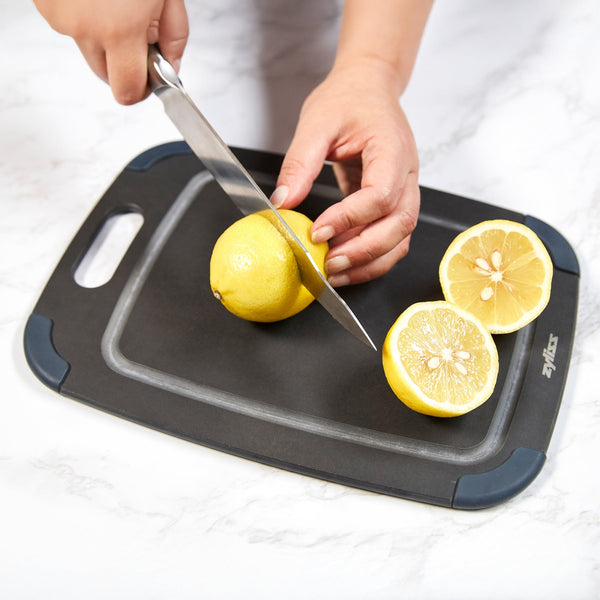A person slices a lemon on the Zyliss Wood Fibre Cutting Board, made from recycled wood fibres, set on a white marble surface. One lemon is being cut while two lemon halves rest on the board. #Small