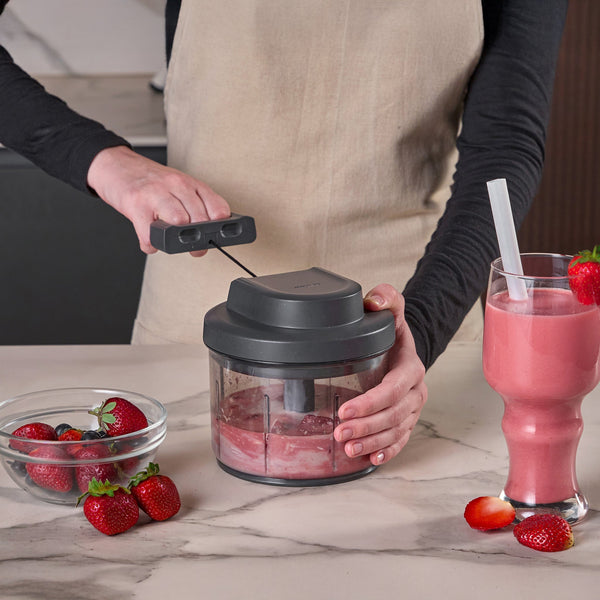 Wearing a beige apron, a person uses the Zyliss Easy Pull Food Processor to blend strawberries on a marble counter. Fresh berries are in a bowl nearby, along with a tall glass of strawberry smoothie with a straw.
