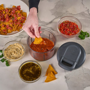 A hand uses the Zyliss Easy Pull Food Processor to make fresh salsa, then dips a tortilla chip into a bowl of salsa surrounded by guacamole, hummus, and tomato salsa, with nachos on a marble surface.
