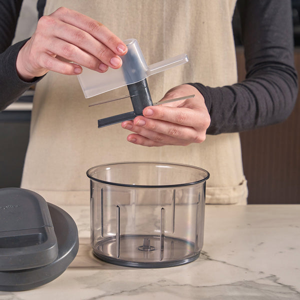 A person assembles the blade and lid of the Zyliss Easy Pull Food Processor over its transparent container on a marble countertop.