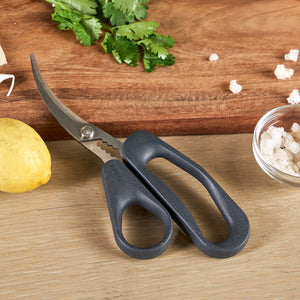 A pair of Zyliss Seafood Scissors with black handles rests on a wooden surface beside a lemon, coarse salt in a small bowl, chopped herbs, and a wooden cutting board.