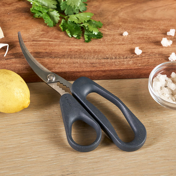 A pair of Zyliss Seafood Scissors with black handles rests on a wooden surface beside a lemon, coarse salt in a small bowl, chopped herbs, and a wooden cutting board.