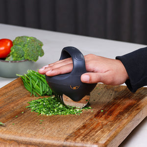 A hand uses the Zyliss Fast Cut Herb Tool, featuring stainless steel cutting wheels, to dice green herbs on a wooden cutting board. Fresh tomatoes and broccoli are visible in a bowl in the background.