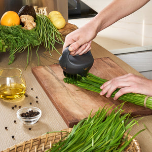 Using the Zyliss Fast Cut Herb Tool with stainless steel blades, someone chops fresh green herbs on a wooden cutting board, surrounded by assorted vegetables, herbs, a glass of oil, and a bowl of peppercorns.