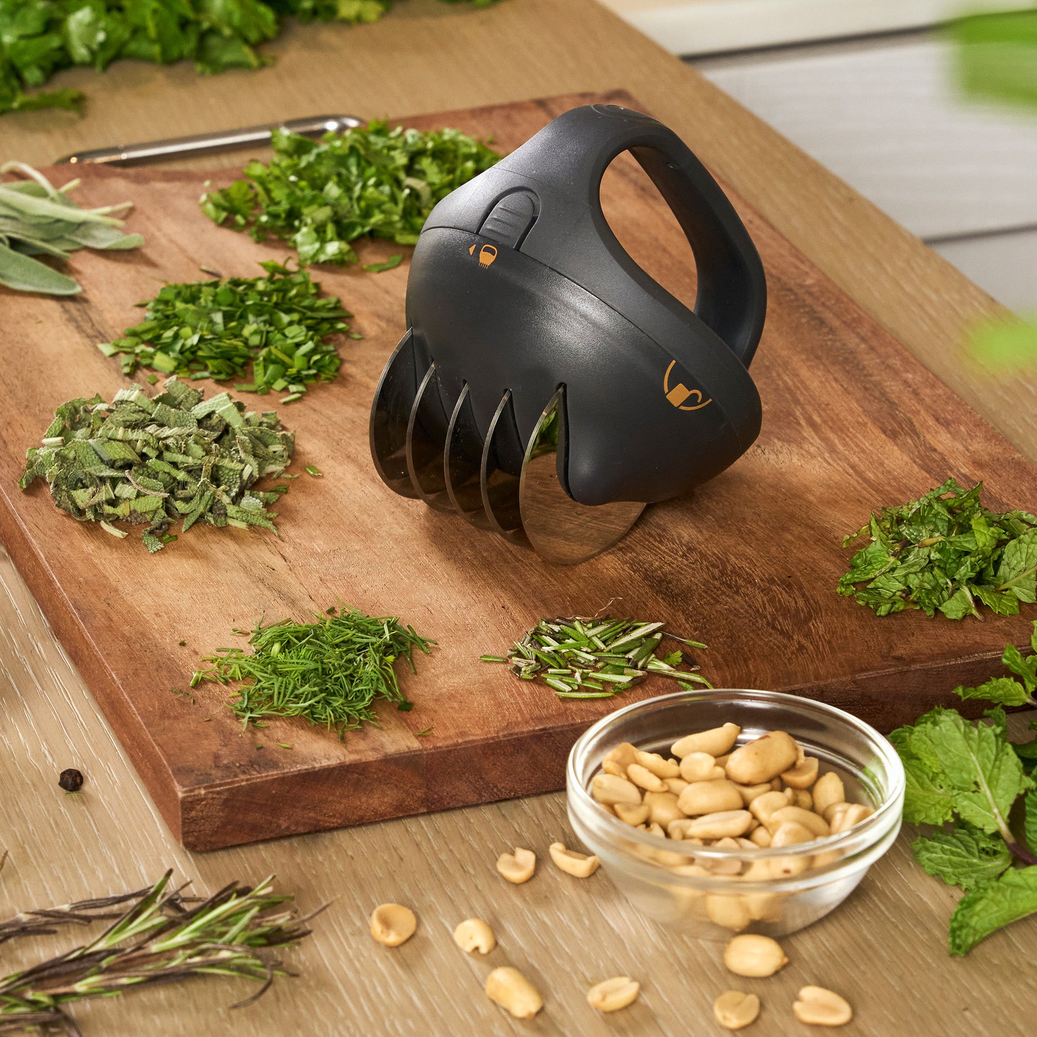 The Zyliss Fast Cut Herb Tool, featuring stainless steel blades, rests on a wooden board amid chopped herbs, fresh leaves, a bowl of peanuts, and a metal herb stripper in a cozy kitchen setting.