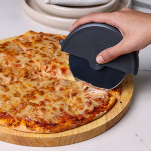 A hand slices a cheese pizza with crispy base using the Zyliss pizza lovers, bundle & save 25% wheel on a wooden board. Stacked white plates sit in the background on a marble surface.