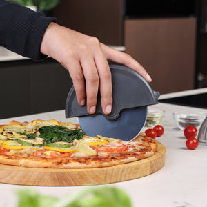 A hand slices a vegetable pizza on a wooden board using the Zyliss Pizza Wheel. Cherry tomatoes and small ingredient bowls sit on the countertop nearby.