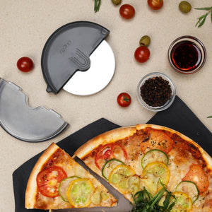 A pizza with tomato and zucchini sits on a dark napkin next to the Zyliss Pizza Wheel, featuring an ergonomic handle and stainless steel blade, surrounded by cherry tomatoes, olives, peppercorns, and a red drink on a beige surface.