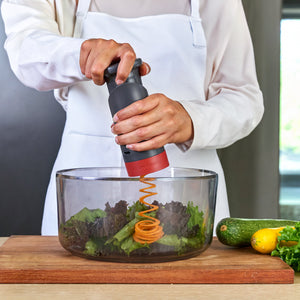 A person uses a Zyliss Vegetable Spiralizer with Reversible Blade and Food Pusher to create long, thin ribbons of carrot into a clear mixing bowl containing salad greens.