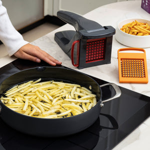 A person stands by a stove frying sliced potatoes as the Zyliss Potato & Vegetable Chipper, with stainless steel blades, a grater, and a bowl of cooked fries rest on the counter; the chipper is dishwasher safe.