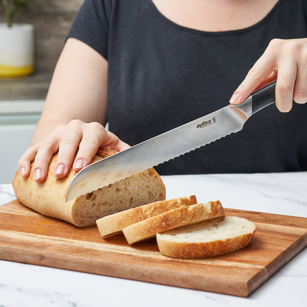 A person slices a loaf of bread on a wooden board using the Zyliss Comfort Pro Bread Knife 20cm / 8, made from German stainless steel with an ergonomic handle. Several fresh slices are neatly arranged on the board. 