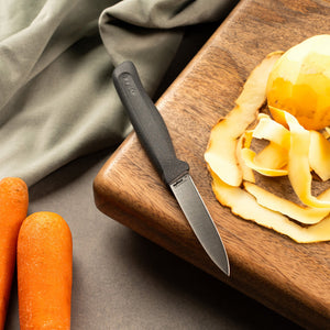 The Zyliss Paring Knife 9cm / 3½ with an eco-friendly handle rests on a wooden cutting board next to a peeled potato, potato skins, two unpeeled carrots, and a green cloth. 