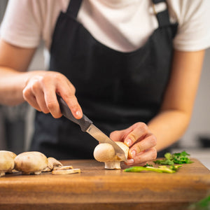 A person in a white shirt and black apron precisely slices a mushroom with the Zyliss Vegetable Knife 9cm / 3½", which has an eco-friendly handle, on a wooden board alongside more mushrooms and fresh herbs.