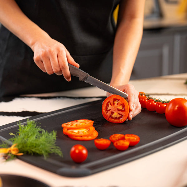 A person is slicing a tomato with the Zyliss Tomato Knife Serrated 11cm (4½") on a black cutting board in a kitchen, surrounded by tomato slices, cherry tomatoes, and fresh herbs.
