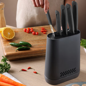 A person pulls a black-handled knife from a Zyliss Universal Knife Block with a flexible bristle insert on the kitchen counter, near a cutting board with sliced tomatoes, whole jalapeños, and a halved orange. Herbs and chili peppers are also on the counter.