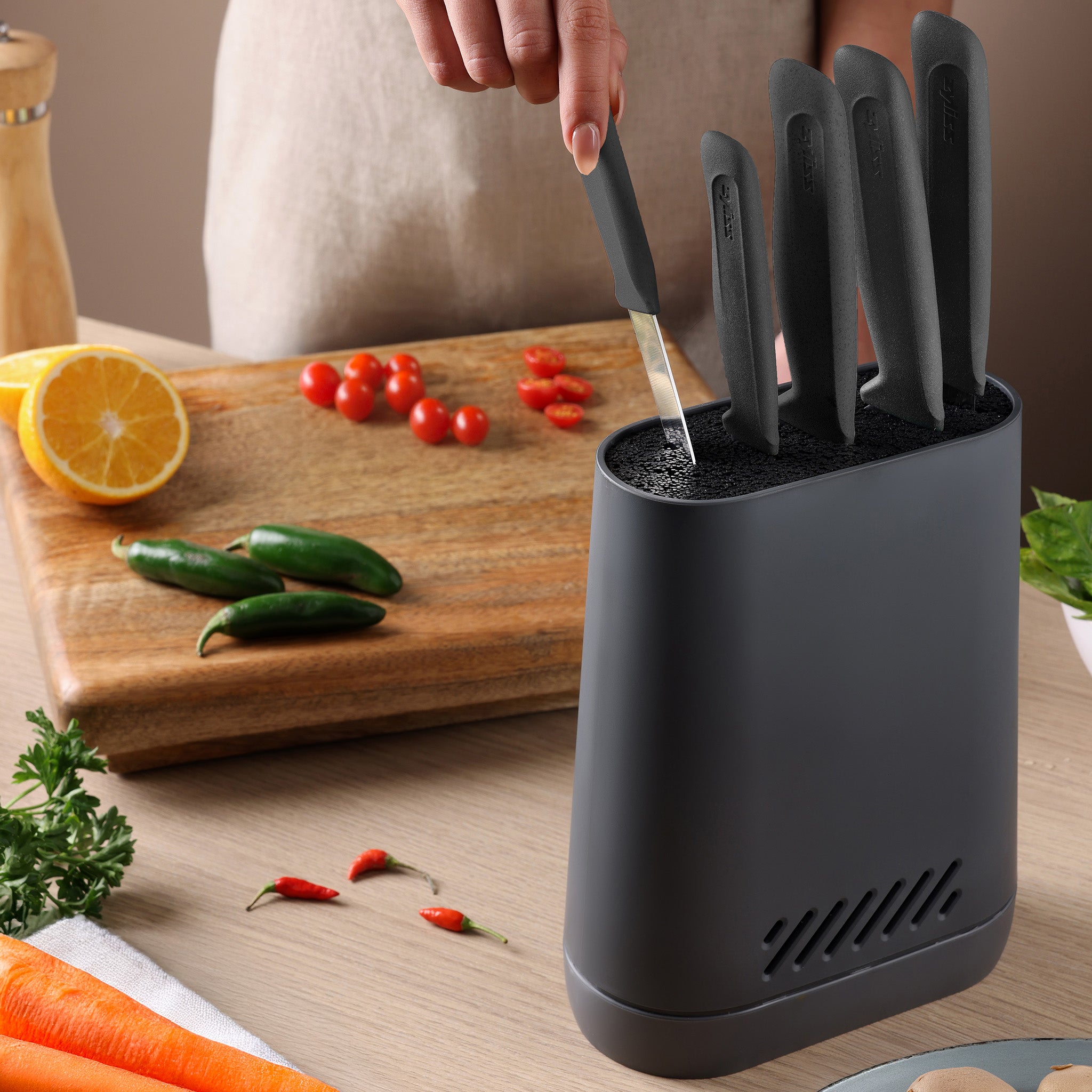 A person pulls a black-handled knife from a Zyliss Universal Knife Block with a flexible bristle insert on the kitchen counter, near a cutting board with sliced tomatoes, whole jalapeños, and a halved orange. Herbs and chili peppers are also on the counter.