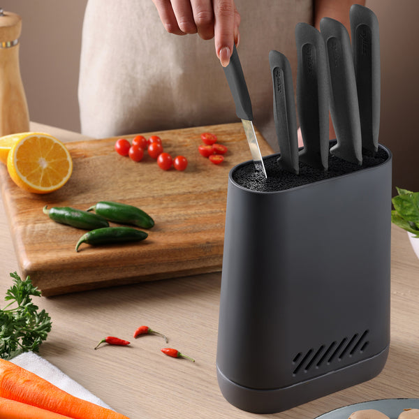 A person pulls a black-handled knife from a Zyliss Universal Knife Block with a flexible bristle insert on the kitchen counter, near a cutting board with sliced tomatoes, whole jalapeños, and a halved orange. Herbs and chili peppers are also on the counter.