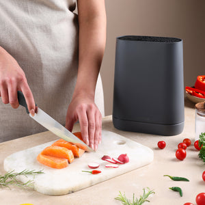 Wearing a beige apron, a person slices raw salmon on a marble board with herbs and veggies nearby, while a Zyliss Universal Knife Block with a flexible bristle insert sits close at hand.