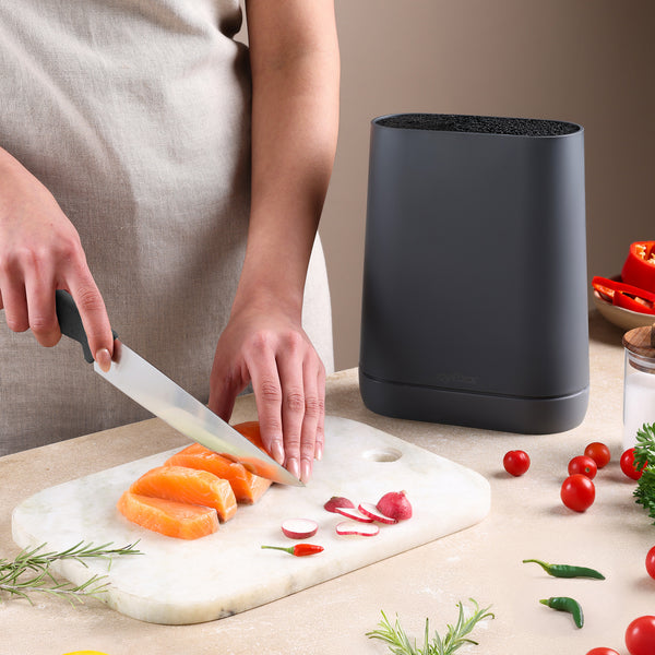 Wearing a beige apron, a person slices raw salmon on a marble board with herbs and veggies nearby, while a Zyliss Universal Knife Block with a flexible bristle insert sits close at hand.