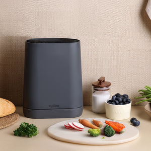 A modern black Zyliss Universal Knife Block with a flexible bristle insert sits on the counter beside bread, sugar, blueberries, green beans, and a round cutting board with radishes, carrots, broccoli, and bell peppers.