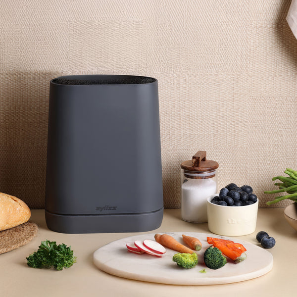 A modern black Zyliss Universal Knife Block with a flexible bristle insert sits on the counter beside bread, sugar, blueberries, green beans, and a round cutting board with radishes, carrots, broccoli, and bell peppers.